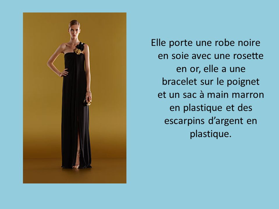 Elle porte une robe noire en soie avec une rosette en or, elle a une bracelet sur le poignet et un sac à main marron en plastique et des escarpins d’argent en plastique.